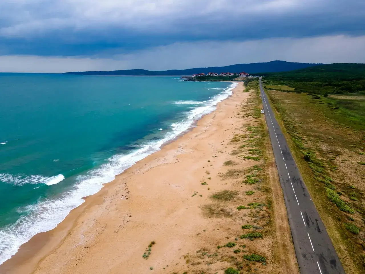 Słoneczny brzeg z turkusową wodą, piaszczystą plażą i drogą wzdłuż wybrzeża. W oddali widać zabudowania i góry.