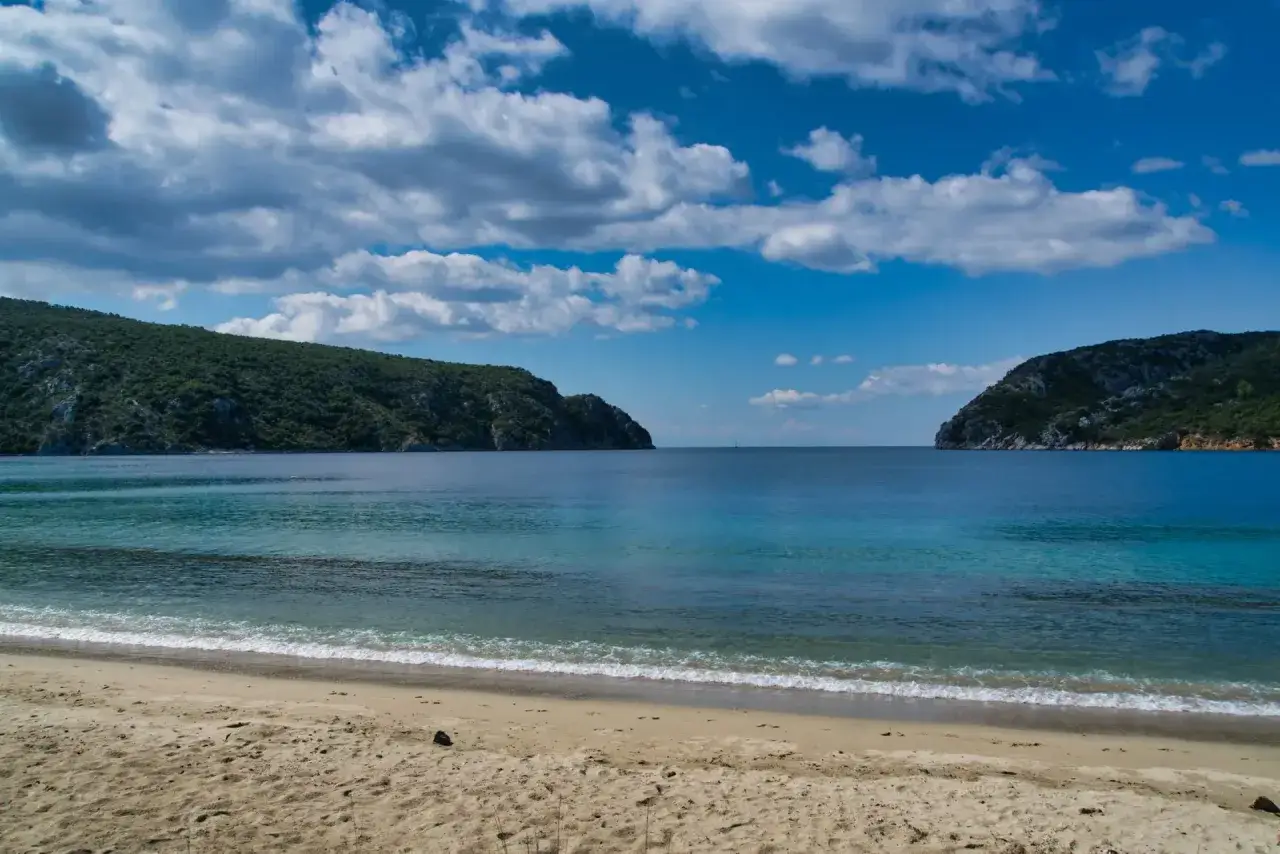 Piękna pogoda w Grecji we wrześniu: piaszczysta plaża, turkusowe morze i zielone wzgórza pod błękitnym niebem z chmurami.